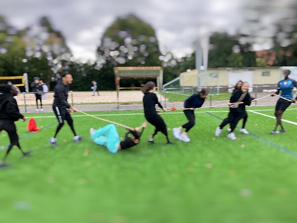 Together in motion – Bewegung, Spaß und Teamgeist Acht Personen spielen auf einem Kunstrasenplatz Tauziehen. Eine Person liegt auf dem Boden, während drei auf der einen und vier auf der anderen Seite am Seil ziehen. Im Hintergrund sind weitere Personen und ein Fußballtor zu sehen. Das Bild ist unscharf.