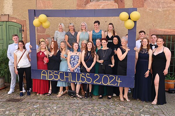 „The Greatest Show“ Zirkus zur Abschlussfeier Eine Gruppe von Personen steht hinter einem großen Rahmen mit der Aufschrift “ABSCHLUSS 2024”. Der Rahmen ist mit gelben Luftballons dekoriert. Die Personen tragen festliche Kleidung und die Szene scheint im Freien vor einem alten Backsteingebäude stattzufinden, was auf eine Abschlussfeier im Jahr 2024 hinweist.