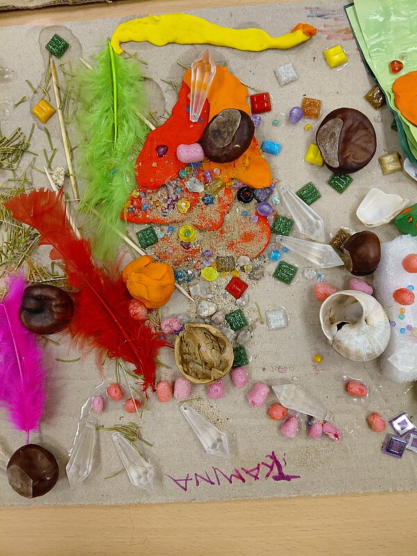 Bunte Materialcollage auf Karton mit dem Namen „TAMINA“: farbige Federn, Kastanien, Muscheln, Walnussschale, Perlen, Sand, kleine Glaskristalle und Knetmasse bilden ein lebhaftes, strukturreiches Muster.
