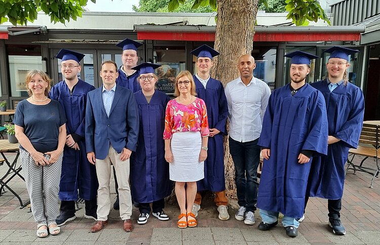Gelungene Zeugnisfeier Gruppenbild der Absolvent*innen in einem Biergarten unter großen Bäumen. Ein Teil der Personen ist in dunkelblaue Talare gehüllt und trägt Absolventenhüte.