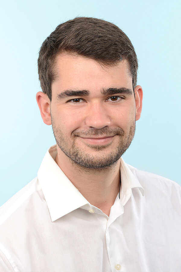 Alumni im Gespräch: Interview mit Felix Täumer Passbild des Absolventen Felix Taeumer, der in weißem Hemd, mit dunklen kurzen Haaren, braunen Augen und einem Dreitagebart in die Kamera grinst. Der Hintergrund ist in dezentem Hellblau gehalten.