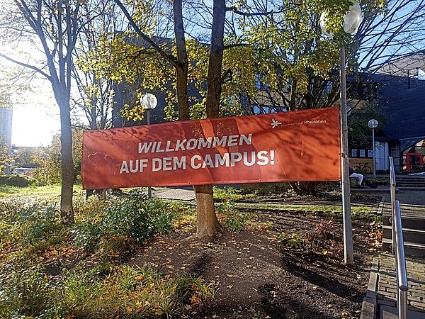 Ein großes rotes Banner mit der Aufschrift Willkommen auf dem Campus ist vor der Hochschule RheinMain gespannt. Die Sonne scheint freundlich an einem Herbsttag.
