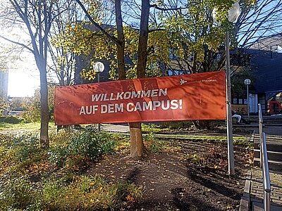 Ein großes rotes Banner mit der Aufschrift Willkommen auf dem Campus ist vor der Hochschule RheinMain gespannt. Die Sonne scheint freundlich an einem Herbsttag.