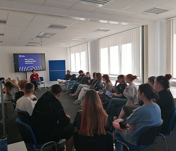 Projekt „Peer-Ausbildung: Schulkino“ durch Alter Gasometer e.V. Zwickau Teilnehmende sitzen im Stuhlkreis und schauen gemeinsam einen Film oder ein Video, das auf einem großen Bildschirm läuft.