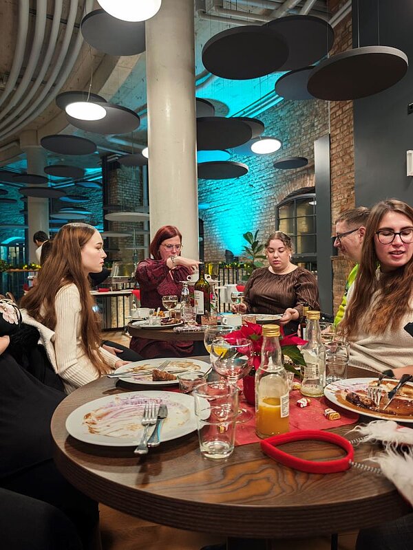 Eine Gruppe von fünf Personen sitzt an einem runden Tisch in einem Restaurant mit industriellem Ambiente, das durch Ziegelsteinwände, moderne Beleuchtung und große runde Lampen an der Decke geprägt ist. Die Personen sind in Gespräche vertieft und essen gemeinsam. Auf dem Tisch stehen Teller mit Essensresten, Gläser, Flaschen mit Getränken, eine Karaffe mit Orangensaft, sowie Besteck und Servietten. Eine Person hält eine Suppenschale hoch. Der Tisch ist mit einer roten Tischdecke dekoriert. Die Atmosphäre wirkt gesellig und entspannt.