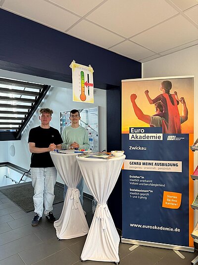 Zwei junge Männer stehen hinter einem Informationstisch der Euro Akademie Zwickau. Der Tisch ist mit Informationsmaterial und einem Roll-up-Banner der Euro Akademie versehen, auf dem „Genau meine Ausbildung“ und die Website „www.euroakademie.de“ zu lesen sind. Im Hintergrund hängt ein Thermometer-Poster mit einer Spendenanzeige.