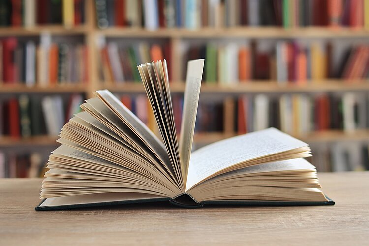 Ein aufgeblättertes Buch liegt auf einem Tisch vor Bücherregalen.