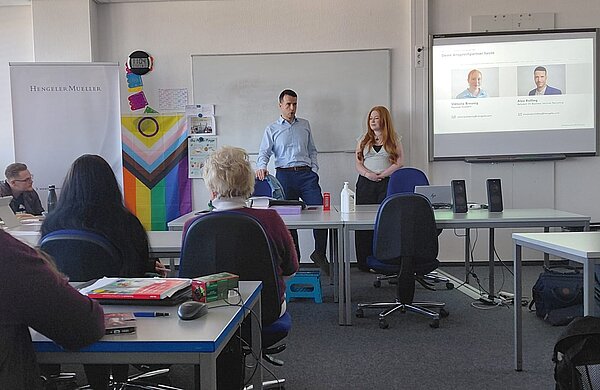 Präsentation im Klassenraum: Zwei Personen stellen sich vor, während auf dem Bildschirm Informationen zu Ansprechpartnern zu sehen sind.
