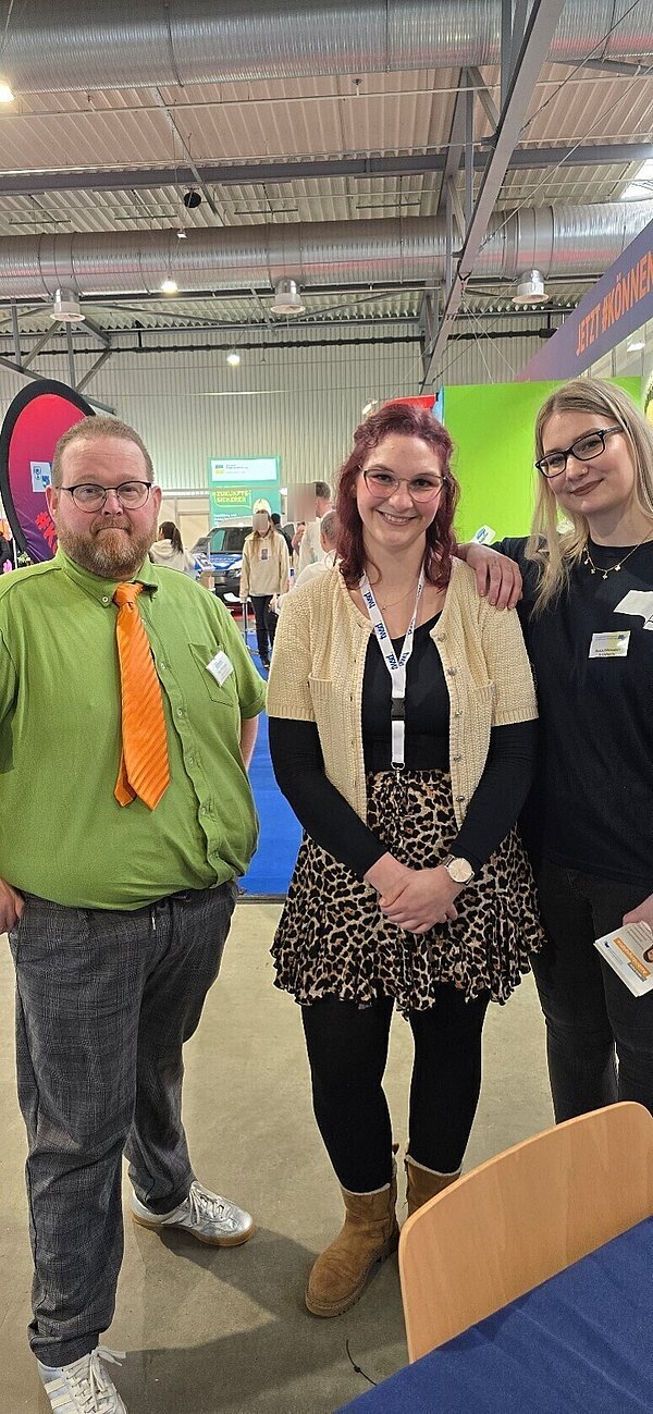 Ein Mann und zwei Frauen stehen lächelnd in einer Messehalle.