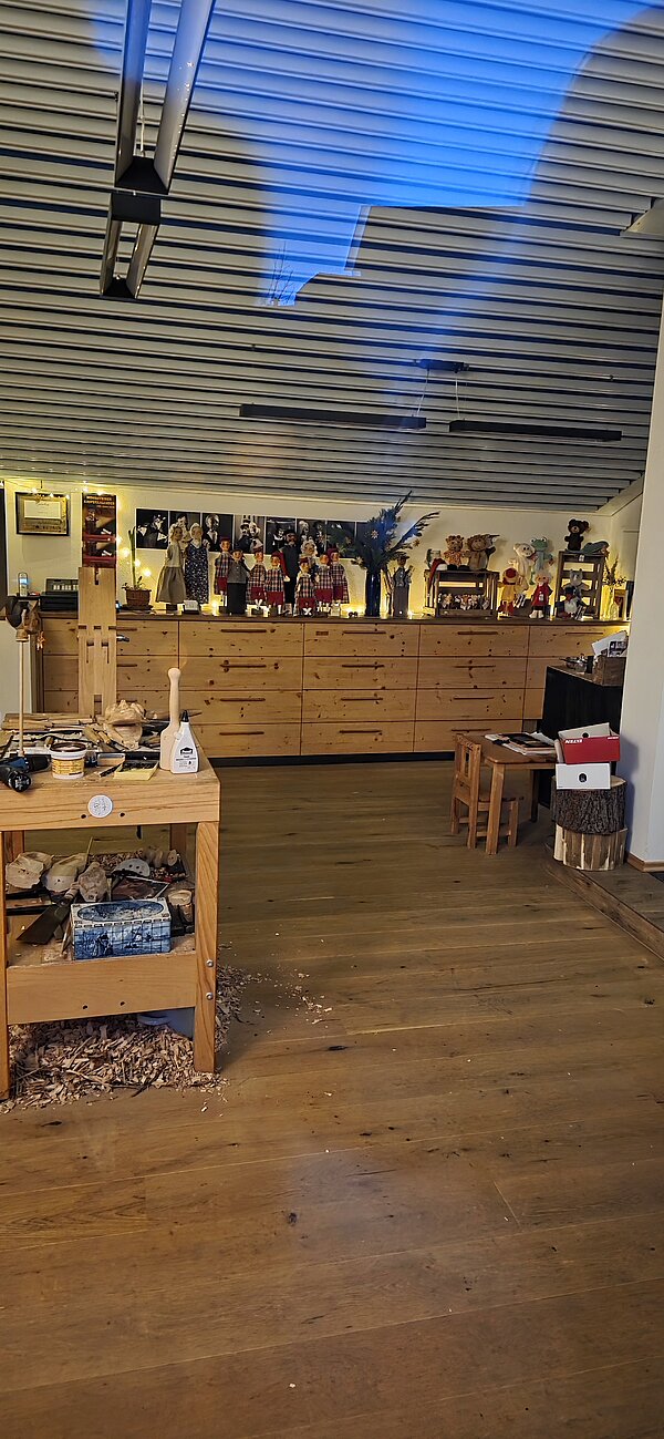 Innenraum einer Holzwerkstatt mit Werkbank, Holzspänen und handgeschnitzten Puppen, die auf einer langen Kommode dekorativ ausgestellt sind.