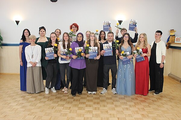Absolventinnen und Absolventen in feierlicher Kleidung sowie Euro Akademie Broschüren in der Hand posieren für ein Gruppenfoto im großen Saal mit Holzparkett.