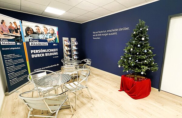 Infobereich der Euro Akademie mit Roll-up-Bannern, Prospektständern, kleinen Bistrotischen mit Metallstühlen und einem dekorierten Weihnachtsbaum vor dunkelblauer Wand.