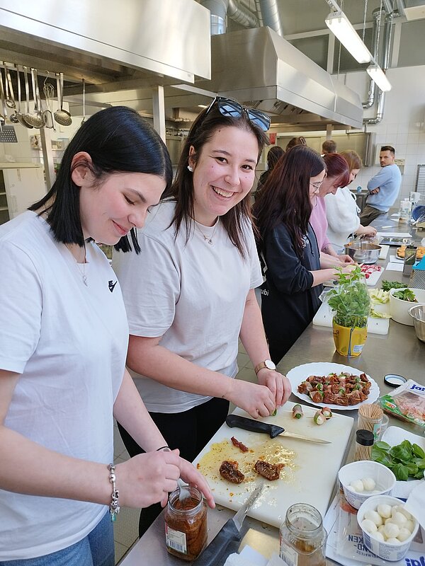 In einer Küche bereiten junge Menschen kleine Häppchen vor; im Vordergrund lachen zwei junge Frauen in die Kamera.