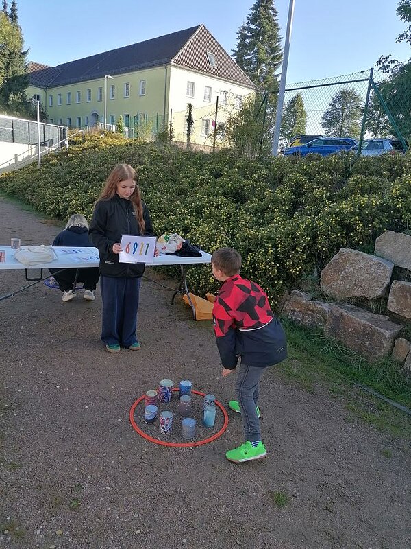 Person mit Schild ‚6911' und ein Junge, der sich über einen Kreis aus orangefarbenen Hütchen mit bunten Dosen beugt – eine Station des Spiel-Sportfests auf einem Kiesweg.