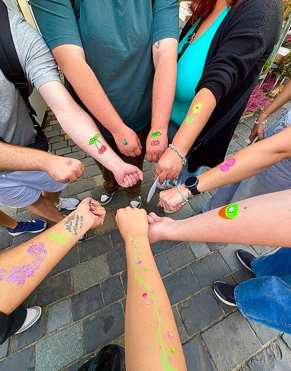 Mehrere ausgestreckte Unterarme zeigen bunte, temporäre Tattoos mit verschiedenen Motiven wie Kirschen, Blumen, Blättern und einem Smiley. Die Tattoos sind auf heller Haut aufgebracht und wirken frisch. Die Personen tragen lässige Kleidung, darunter T-Shirts und Armbänder. Der Hintergrund besteht aus Pflastersteinen und einigen Pflanzen.
