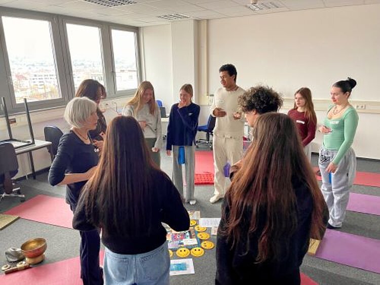 Eine Gruppe von Menschen stehen im Kreis. Einige halten Yogablöcke zwischen den Beinen und halten eine Hand an den Bauch. Die Teilnehmenden machen nach, was ihnen die Übungsleiterin vormacht. Auf dem Boden liegen Yogamatten und Karten.