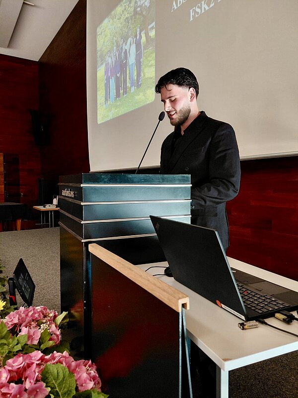 An einem Rednerpult steht ein junger Mann in schwarzem Hemd. Im Hintergrund ist eine Leinwand mit einer Präsentation zu sehen. Im Vordergrund sind ein Tisch mit Laptop sowie rosafarbene Blumen zu sehen.