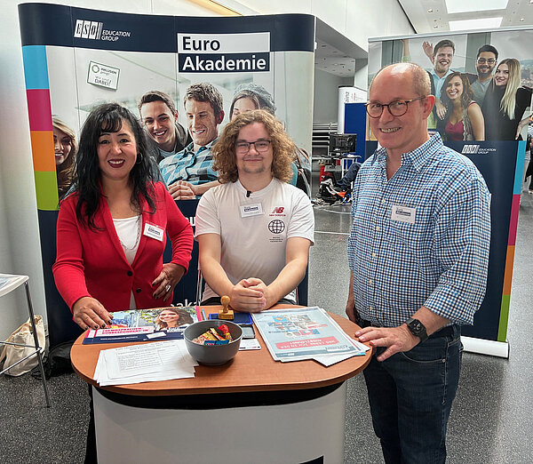Euro Akademie Mainz auf BIM Rheinhessen Zwei Lehrkräfte der Euro Akademie Mainz stehen lächelnd neben einem sitzenden Schüler am Messestand. Der Tisch ist mit Informationsmaterial und einem Snackbehälter ausgestattet.