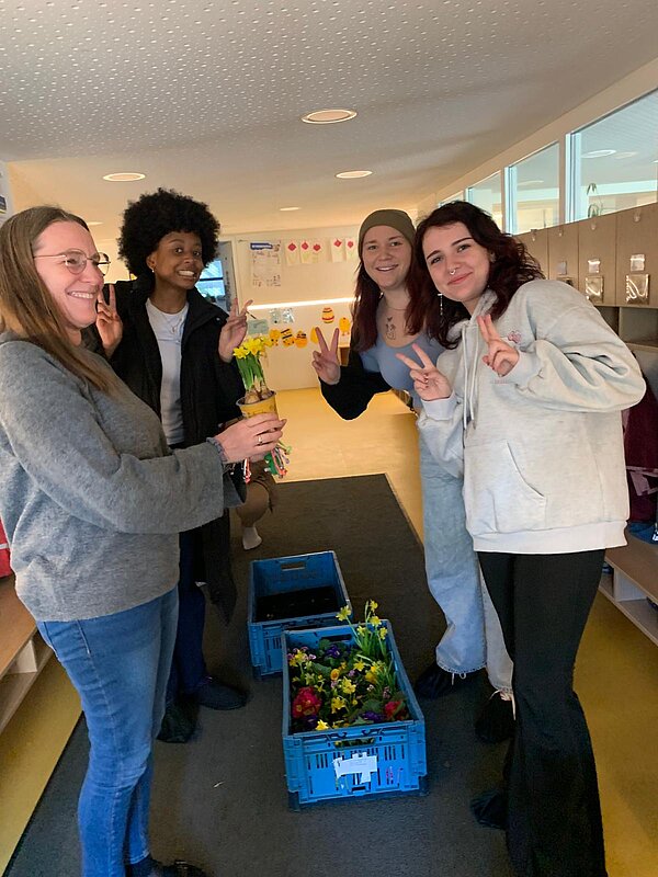 Vier Frauen lächeln in die Kamera; drei davon zeigen Peace-Zeichen, während eine Blumen in der Hand hält; zwischen den Frauen steht eine Kiste mit weiteren Blumen.