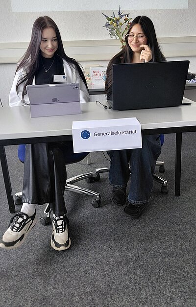 Zwei lächelnde Schülerinnen sitzen mit Laptops an einem Tisch, an dem ein Schild mit der Aufschrift "Generalsekretariat" hängt.