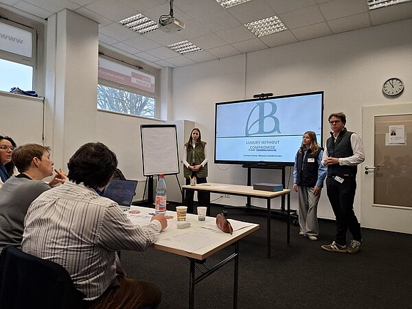 In einem Konferenzraum stehen zwei Personen vorn neben einem großen Bildschirm, auf dem ein Logo mit den Buchstaben 'AB' und der Text 'LUXURY WITHOUT COMPROMISE' sowie weitere Zeilen zu sehen sind. Zwei weitere Personen sitzen an einem Tisch und blicken in Richtung Präsentation. Auf dem Tisch liegen Papiere, Stifte, eine Wasserflasche und Becher. Im Hintergrund befinden sich eine weiße Wand, ein Fenster, eine Uhr und eine geschlossene Tür. Die Atmosphäre wirkt sachlich und konzentriert.