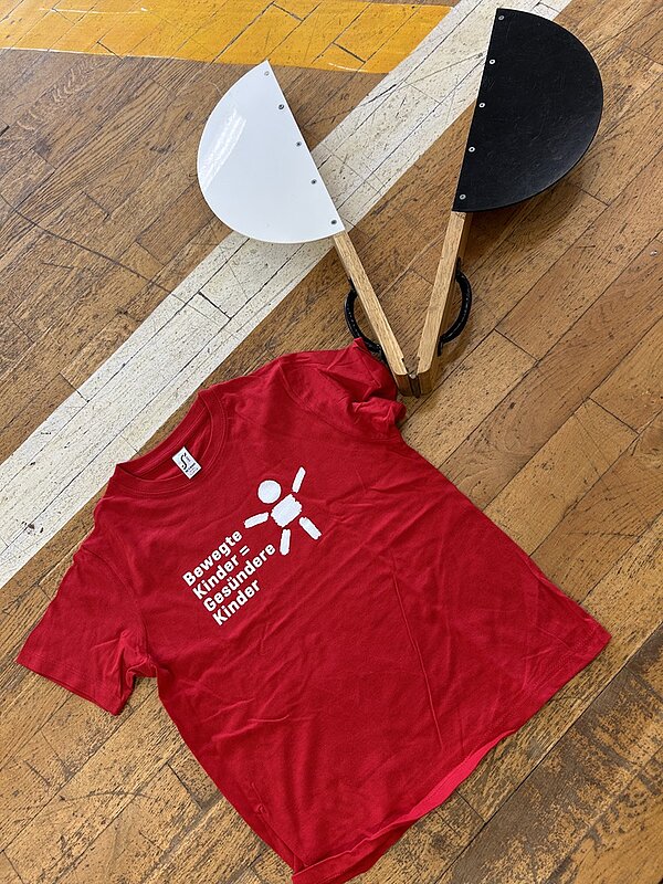 Rotes T-Shirt mit weißem Logo der Initiative Bewegte Kinder = Gesündere Kinder liegt ausgebreitet auf einem Turnhallenboden. Neben dem Shirt befinden sich zwei halbkreisförmige Bewegungs- oder Testgeräte aus Holz mit jeweils einer hellen und einer dunklen Seite, vermutlich Hilfsmittel für den Motorik-Check im Rahmen des Thüringer Bewegungsprogramms für Drittklässler. Das Setting zeigt Materialien, die von angehenden Erzieherinnen und Erziehern der Euro Akademie Pößneck während der Durchführung des Programms genutzt werden.