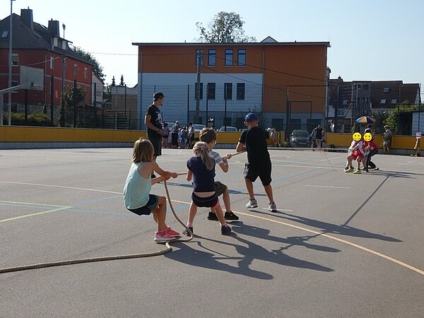 Zwei Gruppen von Kindern stehen drau0en auf dem Sportplatz und treten im Tauziehen gegeneinander an.