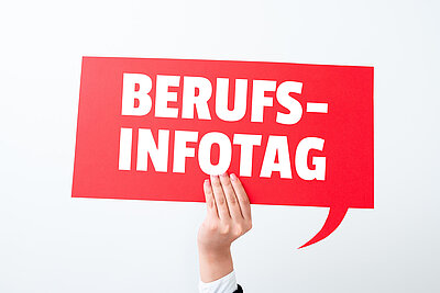 Eine Hand hält ein rotes Schild vor einem hellen Hintergrund, das wie eine rechteckige Sprechblase aussieht, auf der in weißer Schrift "Berufs-Infotag" steht. 