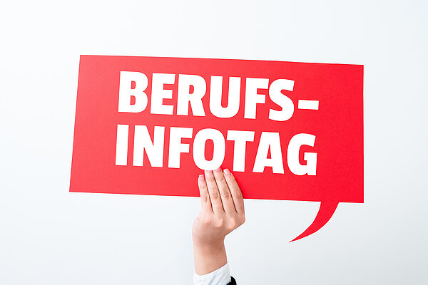 Eine Hand hält ein rotes Schild vor einem hellen Hintergrund, das wie eine rechteckige Sprechblase aussieht, auf der in weißer Schrift "Berufs-Infotag" steht. 