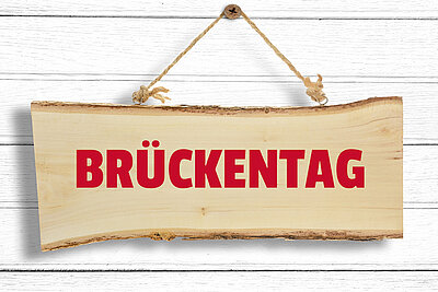  Ein Holzschild mit der Aufschrift "BRÜCKENTAG" hängt an einer weißen Holzwand.