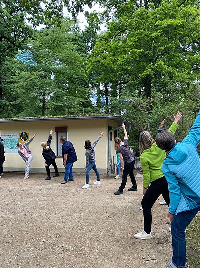 Eine Gruppe von Menschen steht im Freien vor einem kleinen Gebäude und führt eine Übung durch, bei der sie ihre Arme nach oben strecken. Im Hintergrund sind Bäume zu sehen.