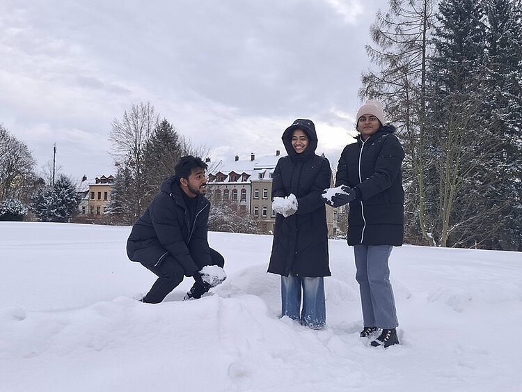 Die Auszubildenden stehen und hocken auf einer schneebedeckten Wiese. Sie tragen dunkle Winterjacken und halten Schnee in den Händen. Im Hintergrund sind mehrstöckige Wohnhäuser mit hellen Fassaden und hohe, kahle Bäume zu sehen. Der Himmel ist bewölkt und die gesamte Szene wirkt winterlich und ruhig.