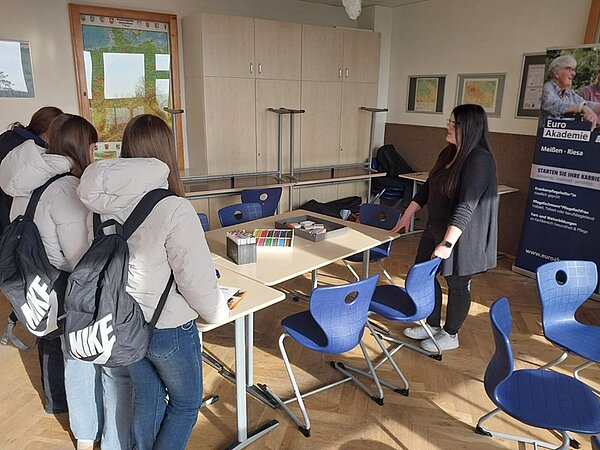 In einem Klassenraum stehen vier Schülerinnen nebeneinander, alle tragen dunkle Jacken und Rucksäcke mit auffälligem Nike-Logo. Sie blicken auf eine Mitarbeitende der Euro Akademie, die vor einem Tisch steht und auf der Informationsveranstaltung Beratungsgespräche mit Interessierten führt. Der Tisch ist mit bunten Karten und Stiften bestückt. Im Hintergrund sind blaue Stühle, ein Schrank, Wandbilder und ein Roll-up mit dem Schriftzug 'Euro Akademie' sowie weiteren Informationen zu sehen.