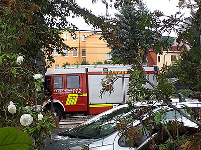 Ein rotes Löschfahrzeug der Feuerwehr mit der Notrufnummer 112 ist teilweise verdeckt hinter Bäumen und parkenden Autos zu sehen.