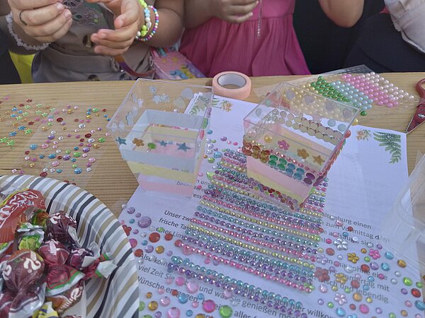 Detailaufnahme gebastelter Plastikbehälter, die mit bunten Glitzersteinen und Perlen beklebt sind, daneben bunte Streudeko und Süßigkeiten.