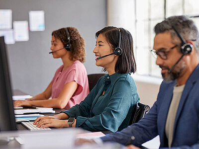 Drei Personen arbeiten in einem Callcenter oder Büro. Alle tragen Headsets und sitzen an Schreibtischen mit Computern. Die Frau in der Mitte, die ein grünes Oberteil trägt, lächelt freundlich und wirkt konzentriert. Links von ihr sitzt eine Frau mit lockigen Haaren und einem rosa Oberteil, die ebenfalls an ihrem Computer arbeitet. Rechts sitzt ein Mann mit Brille und einem dunklen Blazer, der ebenfalls in seinen Bildschirm schaut. Der Raum wirkt professionell und ist mit großen Fenstern ausgestattet, die Tageslicht hereinlassen.