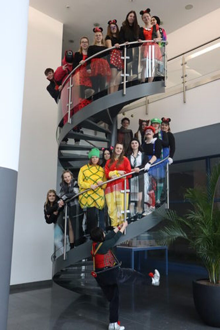 Fasching – Ein Tag voller Spaß und Gemeinschaft Die verkleideten Schüler*innen haben sich auf eine Rolltreppe im Eingangsbereich des Gebäudes für ein Gruppenfoto positioniert.