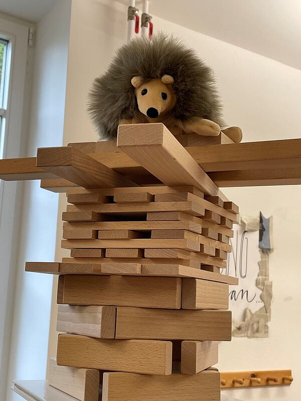 Ein kunstvoll aufgebauter Turm aus Holzklötzen und Brettern. Ganz oben sitzt ein braunes Igel-Stofftier; im Hintergrund eine weiße Pinnwand mit Notizen und eine Garderobe aus Holz.