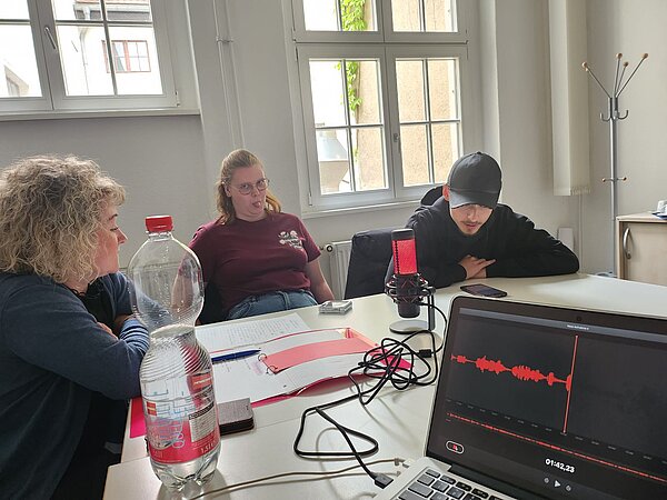 Podcast-Projekt: KAFFEE TRINKEN & SPIELEN Schülerinnen und Schüler der Euro Akademie Halle sitzen an einem Konferenztisch und planen ihr Projekt, vorne sind ein Notebook und Kabel zu sehen