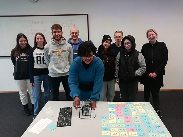 Mit Spaß im Spanisch Unterricht? – Bingo! Lehrkraft dreht an einer kleinen Bingotrommel auf dem Tisch, die Schülerinnen und Schüler stehen hinter ihr.