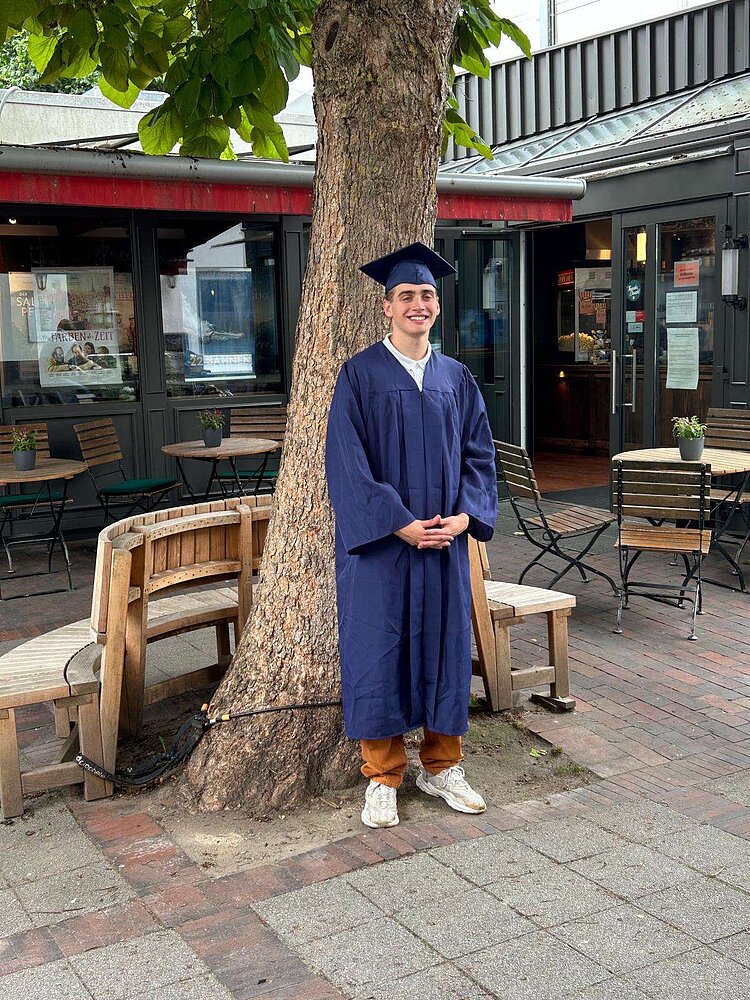 Ein junger Mann mit dunkelblauem Talar und Abschlusshut steht vor einem Baum vor einem Café.