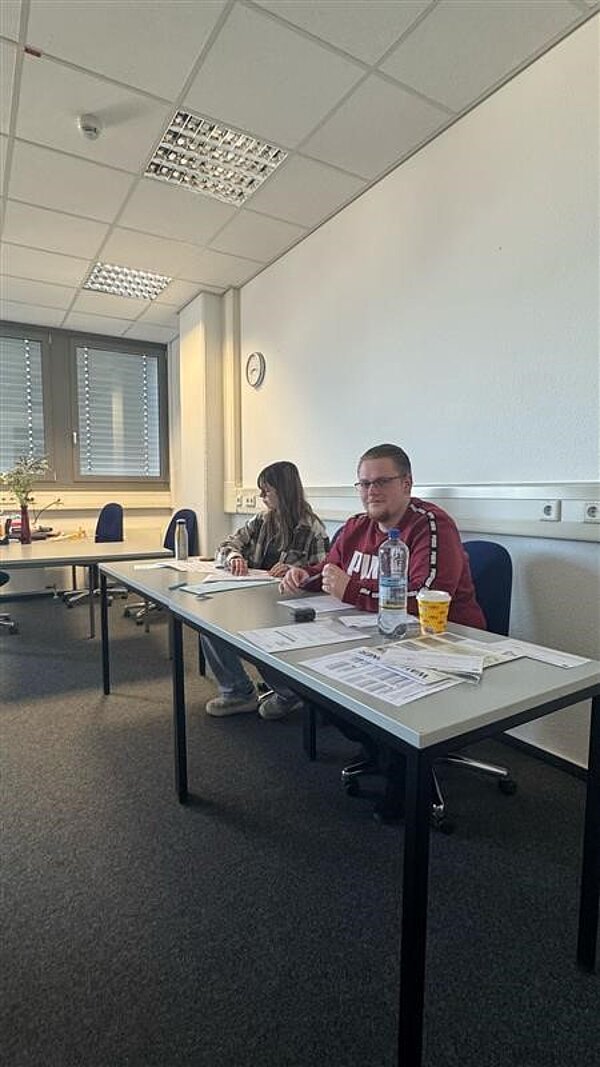 Testwahlen an der Euro Akademie Aschaffenburg am 19. Februar 2025 Zwei Personen sitzen an einem Tisch in einem Wahlbüro. Die Frau links schreibt etwas, während der Mann in einem roten Pullover mit „Puma“-Logo in die Kamera lächelt. Auf dem Tisch liegen Wahlunterlagen und eine Wasserflasche.