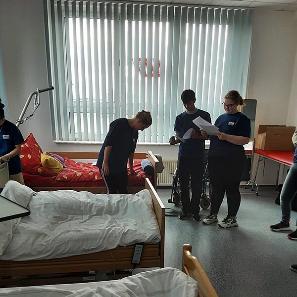 Praxisstunde an der Euro Akademie Bitterfeld-Wolfen Die Auszubildenden stehen in einem Krankenzimmer, mit Betten Triangelgriff. Sie halten Unterlagen in der Hand und tragen blaue Euro Akademie-T-Shirts.