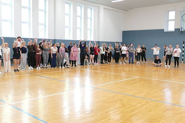 Eine große Gruppe von Menschen steht in einer Reihe in einer Turnhalle. Die Personen sind in lockerer Sportkleidung gekleidet, und einige haben die Arme nach oben gehoben. Die Halle hat große Fenster, durch die Tageslicht einfällt.