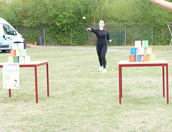 Eine Frau in schwarzer Sportkleidung wirft einen kleinen Ball auf einen Stapel bunter Dosen, die auf einem Tisch stehen. Im Hintergrund sind ein weißer Van und ein Zaun zu sehen. Die Szene findet im Freien auf einem Rasenplatz statt.