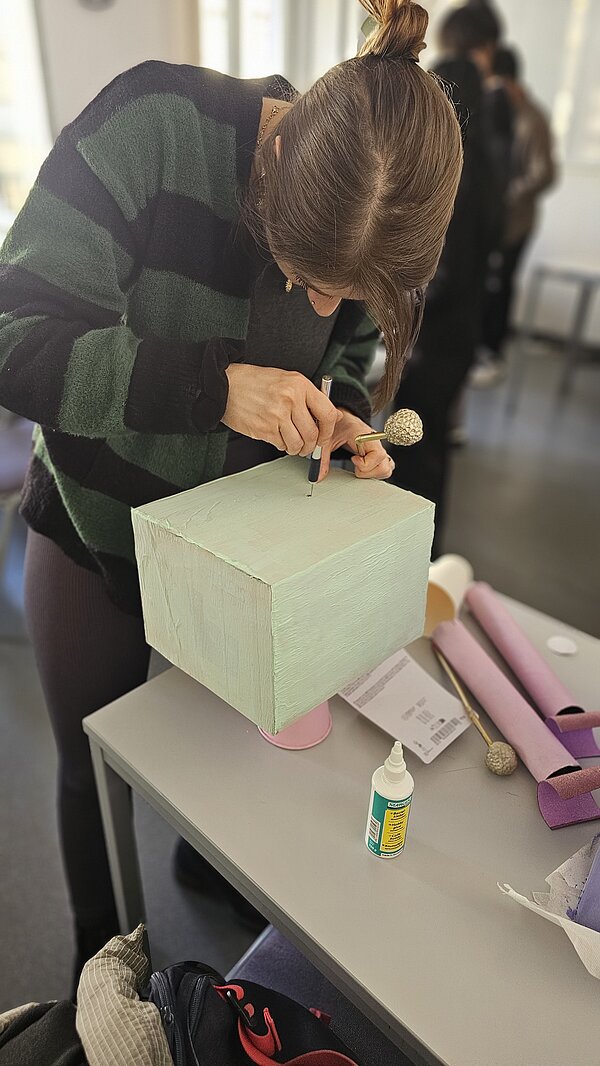 Robotik: Innovative Ansätze in der Pflege Eine Schülerin bohrt ein Loch in die Oberseite eines mittelgroßen, mintfarben bemalten Kartons. In der Hand hält sie eine Art Fühler aus einem goldenen Metallstab mit einer Kugel darauf, der offenbar später in das Loch geklebt werden soll.