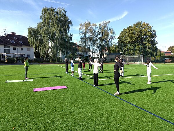 Mehrere Personen stehen in Reihen auf einem grünen Kunstrasenplatz und machen gymnastische Übungen nach, die eine Anleiterin ihnen vormacht. Vor ihnen liegen zwei bunte Gymnastikmatten. Im Hintergrund sind Bäume und ein Gebäude zu sehen.