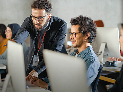 Ein Mann mit Bart und Brille zeigt einem anderen Mann, der ebenfalls eine Brille trägt, etwas auf einem Computerbildschirm. Sie befinden sich in einem modernen Schulungsraum mit weiteren Personen, die an Computern arbeiten.