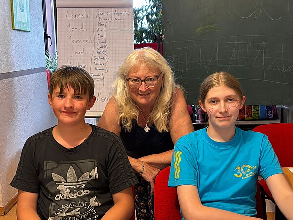 Sprachferien der Euro Akademie Jena und Erfurt: eine 20-jährige Erfolgsgeschichte Ein Schüler und eine Schülerin sitzen auf Stühlen und blicken freundlich in die Kamera. Dahinter eine Lehrerin, die sich zwischen die beiden Kinder platziert hat.