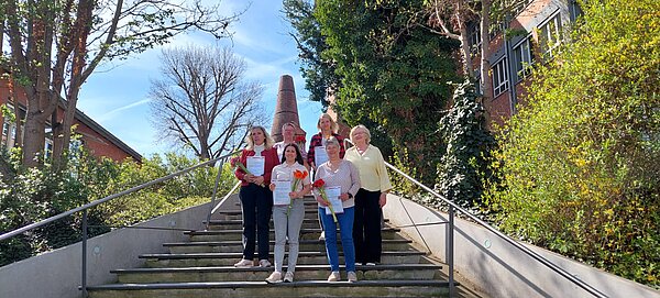 Sechs Frauen stehen auf einer Treppe im Freien, halten Urkunden und Blumen in den Händen und posieren vor Bäumen und Backsteingebäuden.